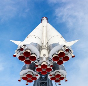 A low-angle shot of a white space rocket with multiple red exhaust nozzles, set against a blue sky with wispy white clouds. The image emphasizes the intricate details of the rocket's engines and structure, conveying a sense of power and technological advancement driven by precise engineering habits.
