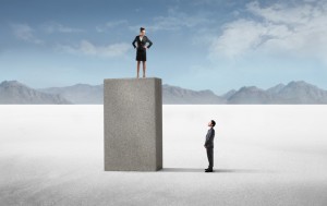 A businesswoman stands confidently on top of a tall concrete block, looking down at a businessman who stands on the ground, gazing up at her. Set against a vast, desolate landscape with mountains in the background and a blue sky overhead, this scene highlights women's rising influence in the corporate world.