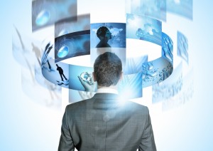 A man in a suit stands facing away, looking at floating digital screens arranged in a semi-circle. The screens display various futuristic images, including silhouettes, a world map, scientific symbols, and abstract designs, all reflecting new technologies. The overall color scheme is blue and white.
