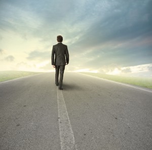A leader in a suit walks alone on a deserted road stretching towards the horizon, under a dramatic sky with clouds. The scene suggests a journey or pathway, with the man seen from behind, moving towards a distant vanishing point.