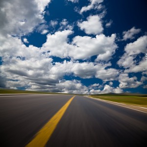 A straight highway stretches under a vibrant blue sky with scattered white clouds. The yellow center line of the road is prominent, guiding travelers towards their goals, while the sides of the road appear blurred, conveying a sense of speed and movement. The surrounding landscape is a mix of green fields.