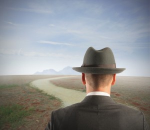 A man in a suit and hat stands facing a winding career path that stretches out into a barren landscape with sparse grass. The path leads toward distant mountains under a clear, blue sky.