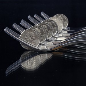 Six forks are placed in a row with the tips facing upwards. Each fork is holding a U.S. quarter in its tines, like a synchronized team of gymnasts. The setup is reflected on a shiny, black surface, creating a mirrored effect of the forks and quarters.