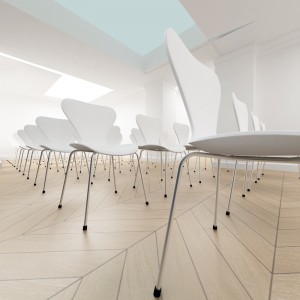 A modern, minimalist meeting room with rows of identical white chairs on light wood flooring, arranged in a semi-circle. Perfect for education sessions, the room has a bright and airy atmosphere with natural light streaming in from a skylight overhead.