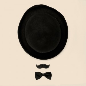 A whimsical arrangement of a black bowler hat, a fake mustache, and a black bow tie on a light beige background evokes the charm of classic dress attire while creating the impression of a face with facial hair and a hat.