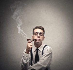 A man wearing glasses, suspenders, and a tie is sitting against a gray backdrop, smoking a pipe. White smoke curls up as he maintains a serious expression, adding an air of performance to the scene.