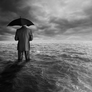 A person in a suit stands knee-deep in an expansive, turbulent sea under a stormy sky, holding an umbrella. The image is in black and white, creating a dramatic and surreal atmosphere that evokes the struggle of unemployment amidst life's storms.