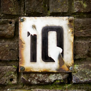 A weathered rectangular metal plate displaying the number "10" is mounted on a textured, moss-covered brick wall. The plate has rust and chipped paint, giving it an aged, rustic appearance akin to an old interview location. Four screws anchor the plate to the wall.