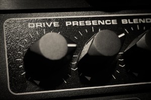 Close-up of a guitar amplifier control panel with three knobs labeled "Drive," "Presence," and "Blend." The knobs are positioned in a dark, textured surroundings, partly illuminated, giving the scene a metallic sheen—almost like the backdrop to an intense interview.