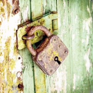 A close-up of an old, rusty padlock securing a weathered green door. The lock and door both show signs of age and wear, with peeling paint on the door and corrosion on the lock. Yes, the surrounding area of the door is also chipped.