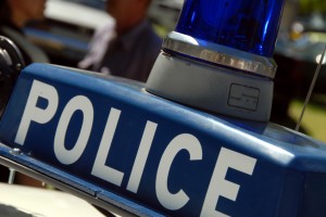 Close-up of a police car light bar with a blue siren and the word "POLICE" in large white letters. The background is slightly blurred, revealing some people and vehicles.