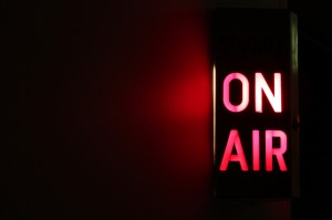 A red and white illuminated "ON AIR" sign is affixed to a dark wall, indicating that a live broadcast or recording is in progress in Studio 1. The room is dimly lit, with the sign casting a red glow on the nearby wall as if resuming an unspoken tradition.