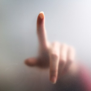 A hand with an extended index finger touches a frosted glass surface. The hand is partly visible and slightly blurred, with the finger's shape discernible through the translucent glass—much like whispered secrets during intimate interviews. The overall tone is soft and muted.