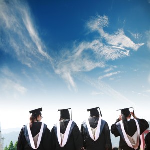 Four MPA graduates in black caps and gowns stand with their backs to the camera, gazing up at a bright blue sky with wispy clouds. Each wears a white stole as they pose in front of scenic, mountainous terrain, ready to embrace the future.