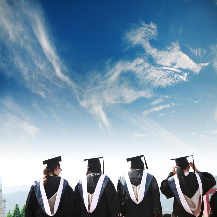 Four MPA graduates in black caps and gowns stand with their backs to the camera, gazing up at a bright blue sky with wispy clouds. Each wears a white stole as they pose in front of scenic, mountainous terrain, ready to embrace the future.