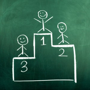 A chalk drawing on a green chalkboard depicts three stick figures on a winners' podium, with the top tier labeled "1" and arms raised in victory. The second and third tiers, labeled "2" and "3," each have a figure standing proudly to receive their award.