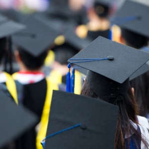 A group of college graduates wearing black caps with blue tassels and gowns are seated during a graduation ceremony. The focus is on the back of a graduate's head in the foreground, with the rest of the graduates and background slightly blurred.