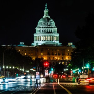 The image shows the illuminated U.S. Capitol Building at night, its dome brightly lit. In the foreground, a busy street, with blurred lights from moving vehicles and traffic signals, suggests a lively urban scene. The setting evokes the bustling environment one might encounter while working a federal job in D.C.