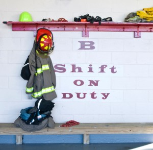 A wall with the text "B Shift on Duty" painted on it. Present above are firefighter gear including a helmet, jacket, and gloves. Additional items are stored on a red shelf, and more clothing rests on a bench below.
