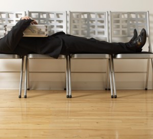 A person in a dark suit is lying down across several interconnected metal chairs in a waiting area, likely resting before an interview. They are holding a light-colored briefcase on their chest. The chairs are against a white wall, and the floor is wooden. The scene suggests the person may be taking a quick nap.