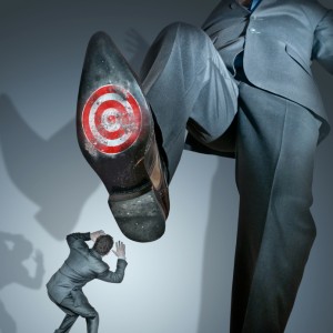 A person dressed in a gray business suit with a target symbol on the sole of a large shoe is stepping on another person in a gray business suit who is crouching and shielding themselves. The scene vividly captures the ruthless nature of office politics, highlighting dramatic contrasts in size and power dynamics.