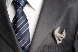 A distinguished leader in a grey pinstripe suit with a blue and white striped tie carries a small adjustable wrench in their suit's breast pocket instead of a pocket square.