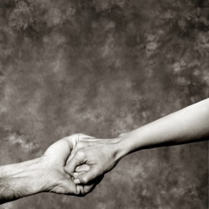A close-up image of two hands against a textured, gray background. An elderly hand, with visible veins and wrinkles, clasps a younger hand gently. The black-and-white photo captures the essence of volunteering, highlighting the deep connection between generations through their touch.