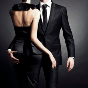 A couple, likely husband and wife, dressed in formal black attire, set against a dark background. The woman is wearing a strapless dress with a ruffled neckline and has her hand on the man's hip. The man is in a black suit with a white shirt and black tie. Both are facing slightly away from the camera.