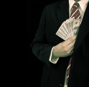 A person wearing a dark suit and a striped tie is holding several US dollar bills in their right hand, partially concealed in their jacket, hinting at potential bribery. The image is set against a dark background.