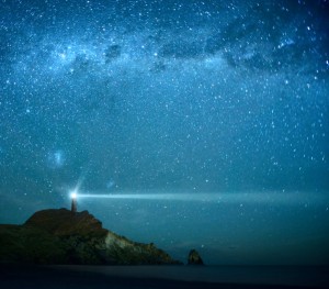 A lighthouse on a rocky cliff shines a bright beam of light across a tranquil ocean under a star-filled night sky, symbolizing success. The Milky Way stretches above in vivid blue tones, adding to the calm and serene scene.