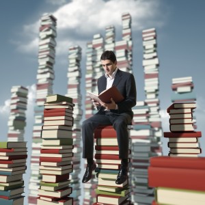 A man in a suit reads a book while sitting on a large stack of books, immersed in research, surrounded by numerous towering book stacks under a partly cloudy sky.