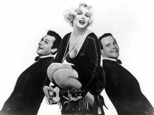 A black-and-white photo shows a woman in a black dress and blonde curly hair kneeling and holding a small guitar. Flanking her on either side are two men, possibly her employers, in suits, leaning back-to-back with her, both smiling and wearing bow ties.