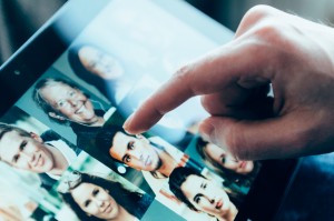 A close-up of a hand touching a tablet screen displaying various profile photos of people. The screen shows a diverse group of faces in a grid layout, indicating a social media or networking application.