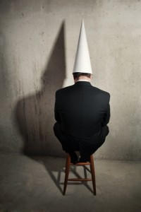 A person sits on a wooden stool facing a concrete wall, dressed in a black suit and wearing a white cone-shaped dunce cap. The lighting casts a large shadow of the hat on the wall, raising questions about their predicament.