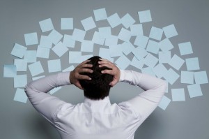 A person in a white dress shirt is facing a gray wall covered with many light blue sticky notes. The person, potentially dealing with project management tasks, is grasping their head with both hands, seemingly overwhelmed or stressed by the number of notes.