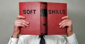 A person in a white shirt and tie is holding a large red book titled "SOFT SKILLS" in bold black letters. The book, held open in front of their face, displays "SOFT" on the left page and "SKILLS" on the right.
