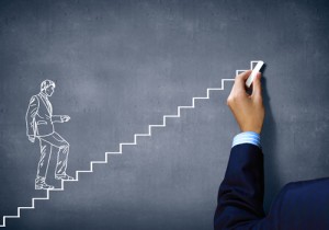 A hand in a suit is drawing white steps on a dark chalkboard. A sketch of a person in a business suit is shown stepping upward on the drawn stairs, symbolizing career progress and achievement.