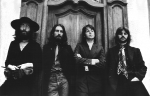 A black and white photo of four men standing in front of an ornate wooden door. From left to right, the first man wears a hat and glasses, the second has long hair and a beard, the third has his arms crossed, and the fourth has a mustache and a patterned scarf. The resolution captures every intricate detail vividly.