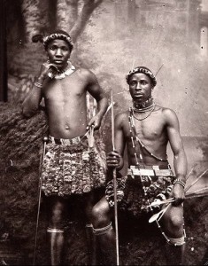 Two men dressed in traditional warrior attire pose for a photograph. They wear beaded ornaments and skirts made of what appears to be animal hide. One man stands holding a spear, and the other is seated with a bow and arrows. The background is an outdoor, natural setting.