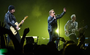 A rock band performs live on stage under green and yellow lights. The lead singer, wearing sunglasses and a leather jacket, sings into the microphone with laser-focused priorities. The guitarist and bassist, clad in dark and stylish clothing, passionately play their instruments.