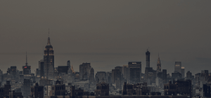 A dimly lit cityscape at dusk featuring numerous skyscrapers. The prominent skyline includes iconic buildings such as the Empire State Building and the Chrysler Building, with construction cranes visible amidst the dense arrangement of structures. The sky is overcast.