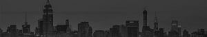A grayscale panorama of a city's skyline at dusk, prominently featuring the Empire State Building on the left. The cityscape is dense with various high-rise buildings and skyscrapers, fading into the distance under a dim, overcast sky.
