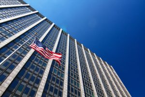 A low-angle view of a tall government building with many windows against a clear blue sky. An American flag is prominently waving in the wind from a flagpole attached to the building.