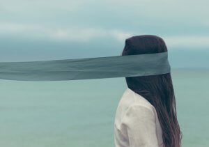 A person with long dark hair stands facing the ocean, with a light-blue ribbon stretching across their eyes, obscuring their vision. The sky is overcast, and the person is wearing a light-colored shirt. The mood is serene and introspective.