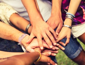 A group of people standing and placing their hands together in the center, stacked on top of each other, symbolizing unity and teamwork. Various skin tones and wrist accessories are visible, showing diversity among the participants.