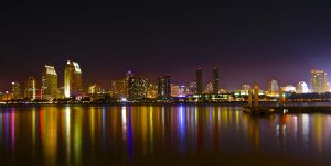 A vibrant cityscape of San Diego is depicted at night, with illuminated skyscrapers reflecting on the calm waters in the foreground. The bright lights create a colorful array on the water's surface, and a dock extends into the water on the right.