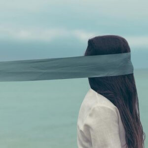 A person with long hair is standing near a body of water with their head partially wrapped in a gray cloth, obscuring their face. The sky is overcast, and the person is wearing a white garment.