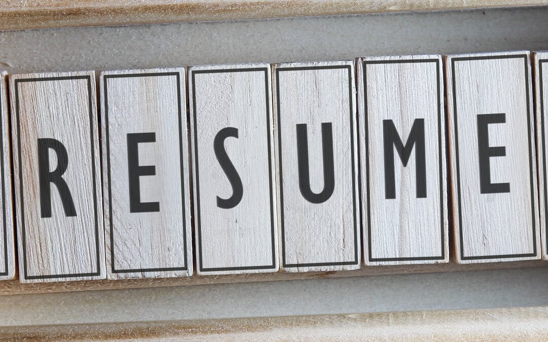 A close-up of wooden blocks arranged in a row, spelling out "RESUME" in black letters. The blocks have a lightly weathered texture, akin to the aged pages of old resumes, and the surrounding background is a muted, textured surface.