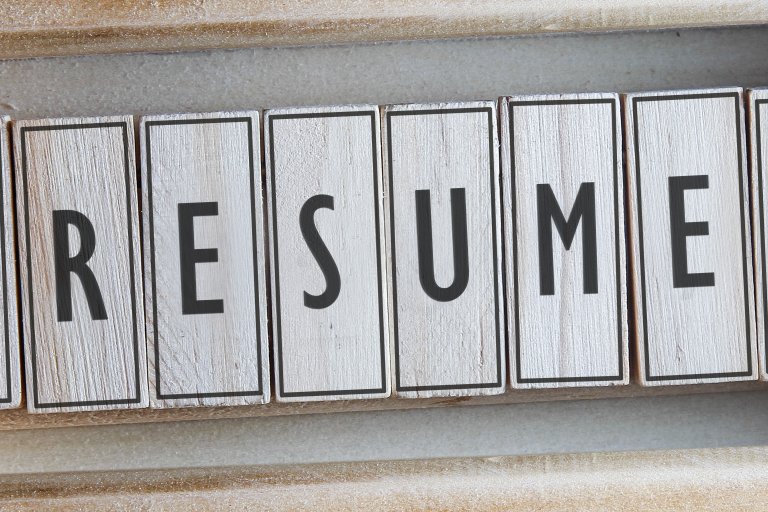 A close-up of wooden blocks arranged in a row, spelling out "RESUME" in black letters. The blocks have a lightly weathered texture, akin to the aged pages of old resumes, and the surrounding background is a muted, textured surface.