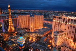 Aerial view of Las Vegas at dusk, featuring brightly lit hotels and casinos. The Eiffel Tower replica and illuminated signs contrast with the darkening sky. Various tall buildings, including "planet" on the facade, create a vibrant and bustling cityscape.